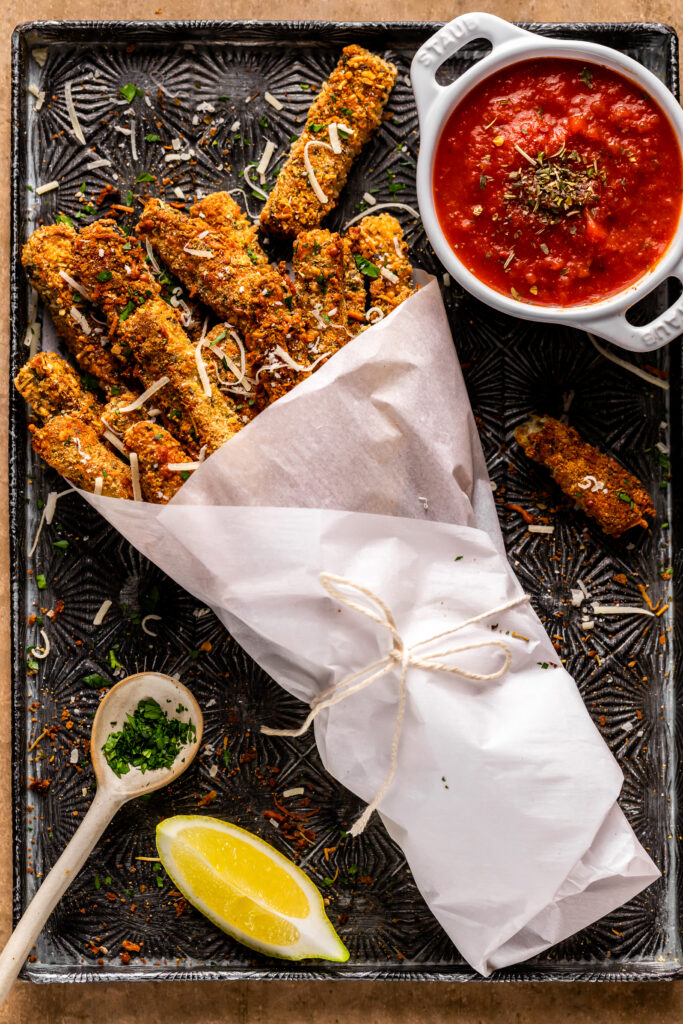 The Eggplant fries are wrapped up in parchment paper with a string tied around the base laying in a baking tray with a side of marinara sauce and a lemon wedge.