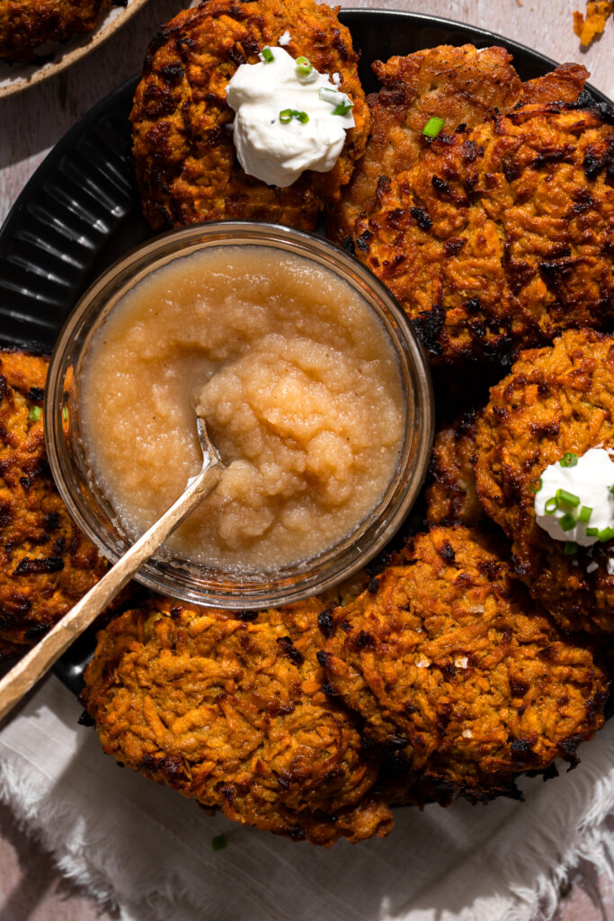 close up of sweet potato latke dish with a spoon dipped into a small bowl in the center of the place filled with apple sauce. Some latkes have a dollop of sour cream and chives sprinkled on top.