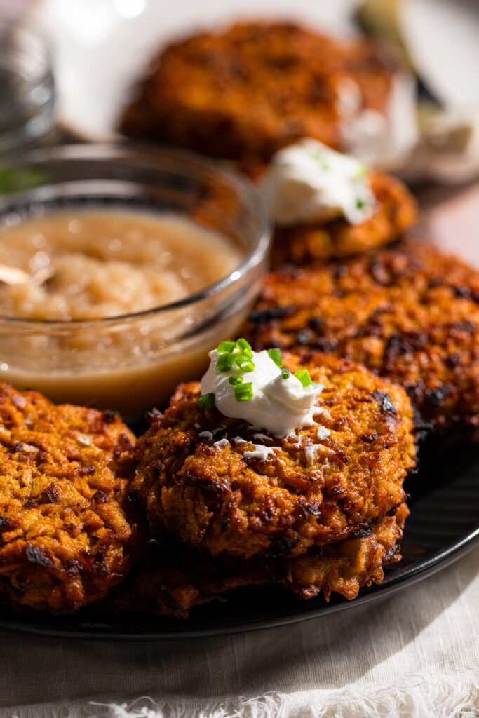 A side profile of the latkes sitting on a black plate with a dollop of sour cream sitting on top. A small bowl of apple sauce sitting at the center of the bowl.