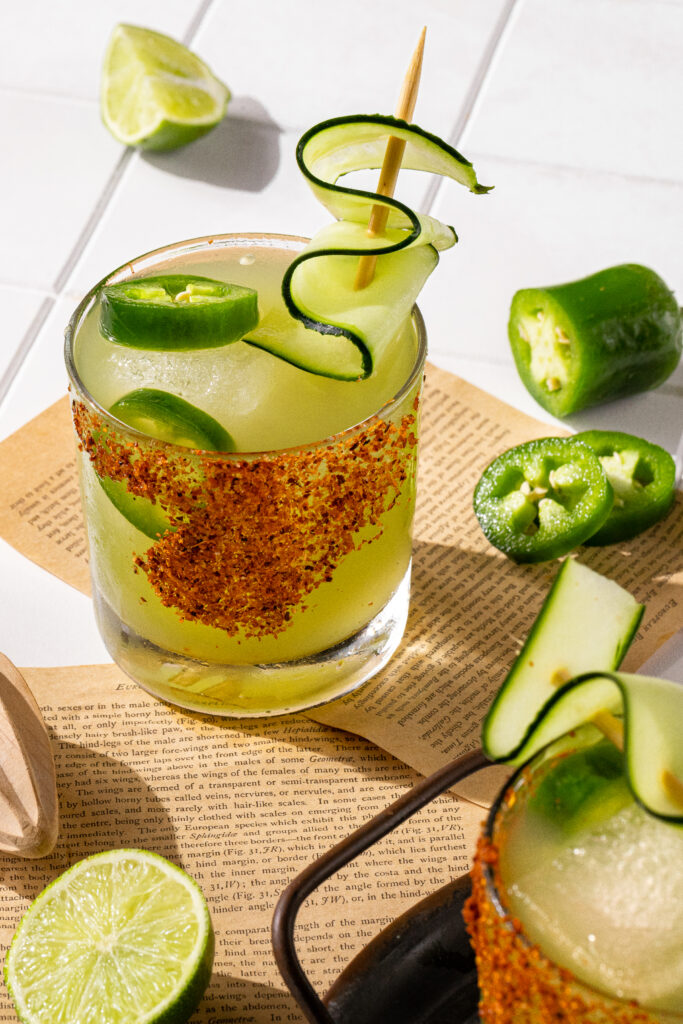 A cucumber margarita ready to serve surrounded by fresh jalapeños and limes.