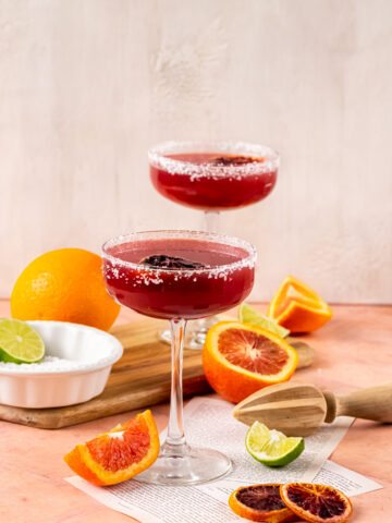 A wide shot of the two margaritas sitting on a table surrounded by slices of blood orange and lime.