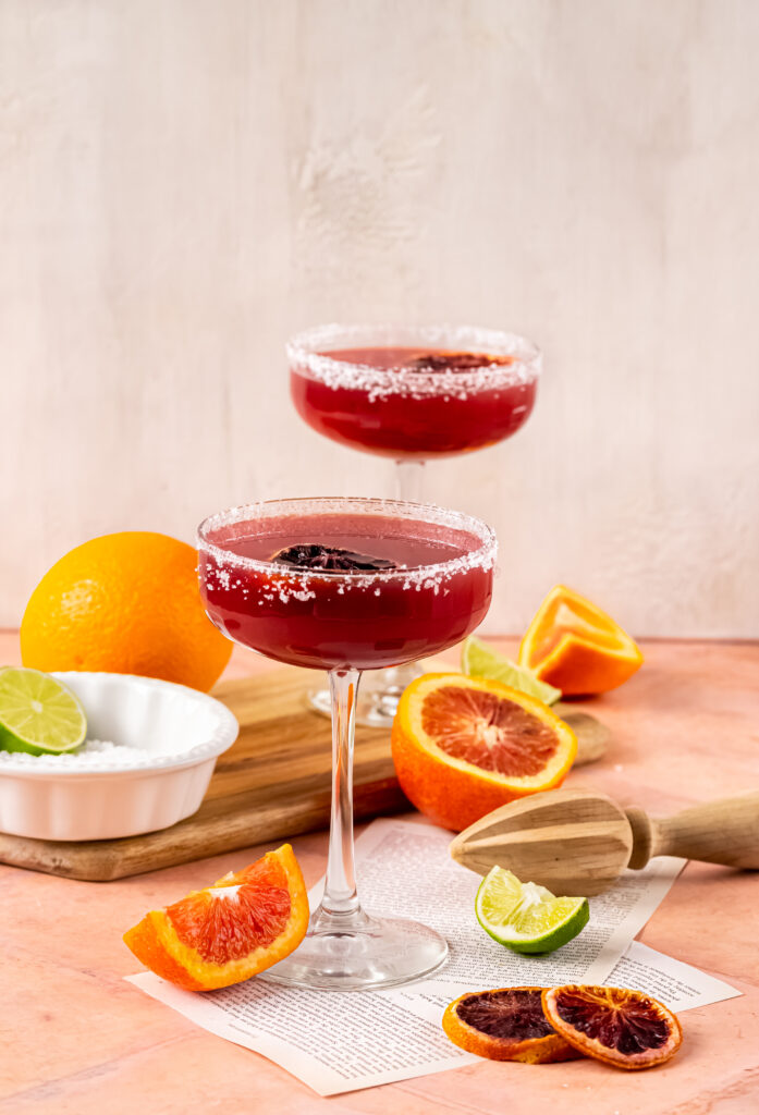 A wide shot of the two margaritas sitting on a table surrounded by slices of blood orange and lime.