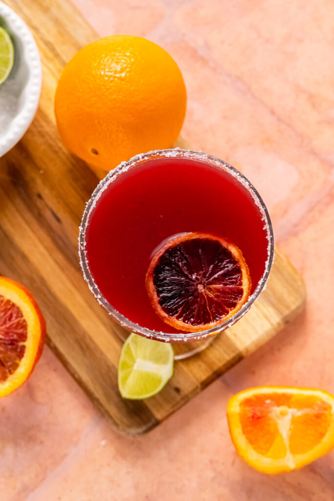 A top view of a blood orange margarita sitting on a cutting board surrounded by blood orange slices and limes.